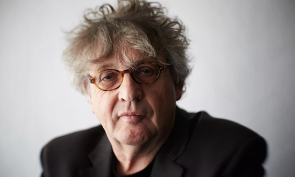 A middle-aged man with tousled hair and round glasses gazes directly at the camera, wearing a black jacket against a plain background.