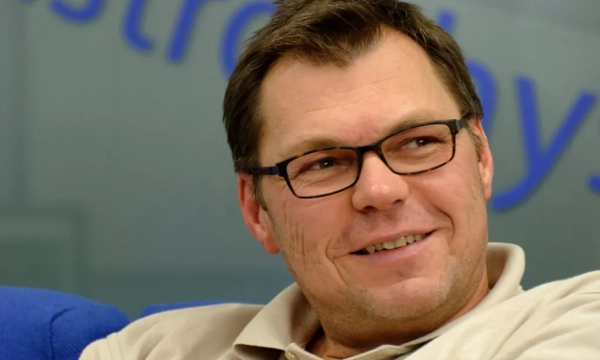 A smiling man with glasses, wearing a beige polo shirt, sitting in a relaxed environment with blue furniture.