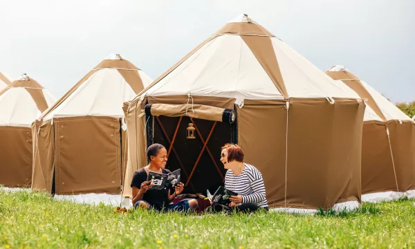 Two women sit on the grass outside brown canvas tents, laughing and reading books, with a cloudy sky in the background.