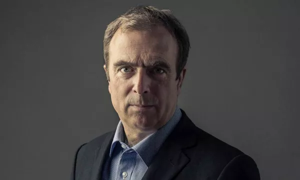 A middle-aged man in formal attire, with short hair, looking intently at the camera against a dark background.