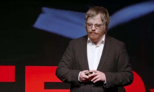 A speaker at a TEDx event, wearing glasses and a dark suit, stands on stage presenting, with a blurred background displaying the TED logo.