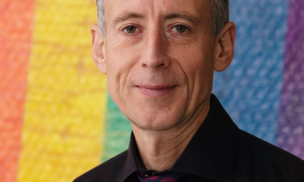 Portrait of a man with gray hair and a black shirt, standing in front of a colorful rainbow background.