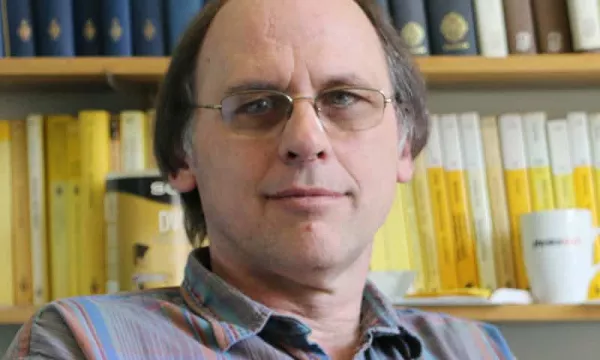 A middle-aged man with glasses and a plaid shirt sitting in an academic office surrounded by books, including various colored volumes on a shelf behind him.
