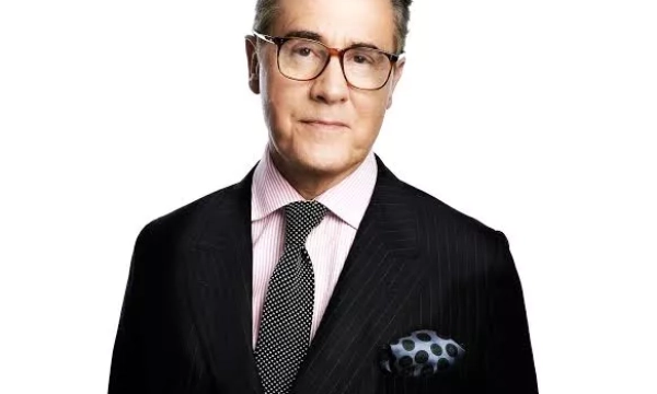 A man in a tailored black suit with a patterned tie and pocket square, wearing glasses, posing against a white background.