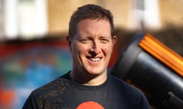 A smiling man wearing a black t-shirt stands outdoors near a large telescope, with colorful graffiti in the background.