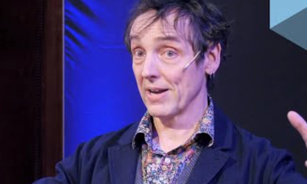A man with tousled hair is speaking passionately, wearing a patterned shirt under a dark jacket, against a blue-lit background.