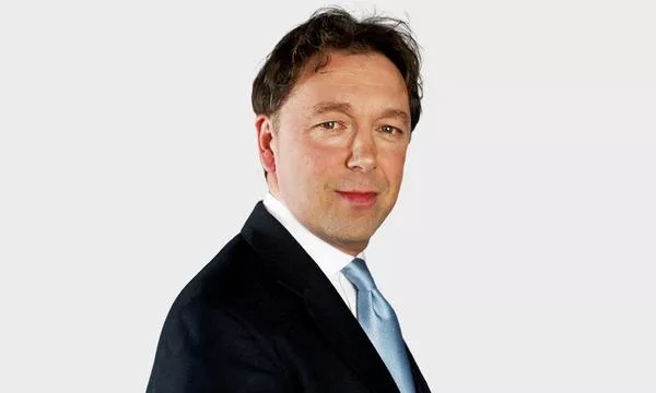 A man in a black suit and light blue tie poses against a plain background, with a confident expression.