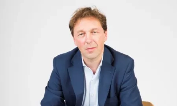 A middle-aged man with medium-length brown hair, wearing a blue suit jacket over a light blue shirt, sitting against a plain white background.