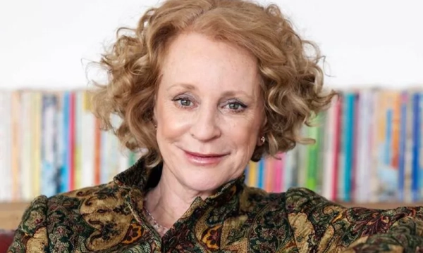 A woman with curly hair smiles warmly, dressed in a patterned jacket, seated against a colorful bookshelf background.