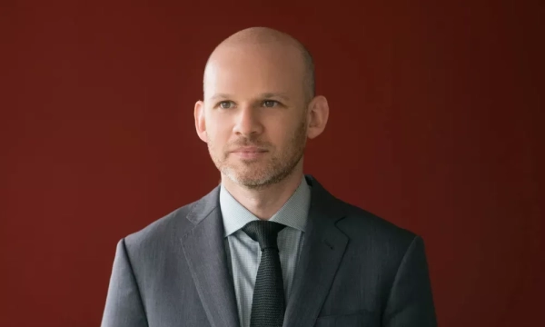 A man with a bald head and beard stands in a dark suit against a red background, looking thoughtfully into the distance.