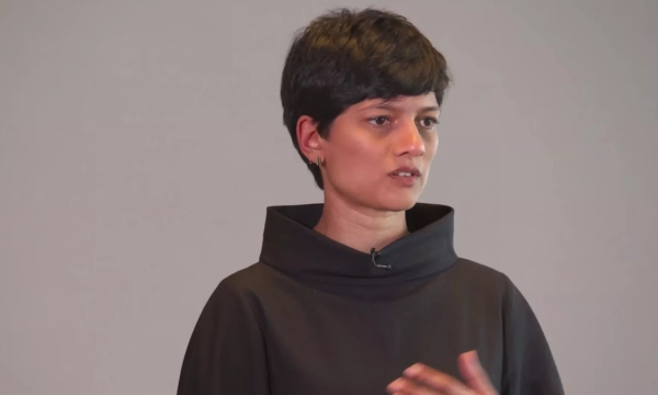 A woman with short black hair wearing a dark, modern outfit speaks thoughtfully during a presentation against a neutral background.