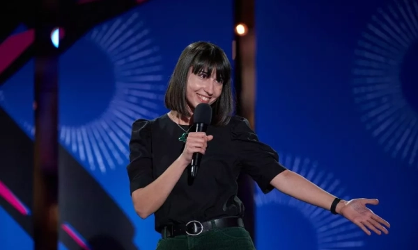 A female comedian in a black top stands on stage, smiling and holding a microphone, with a vibrant blue background featuring geometric designs.