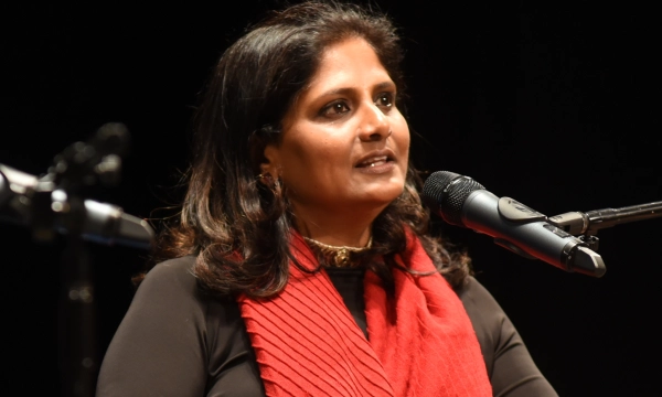 A woman with long dark hair speaks into a microphone, wearing a black top and a red scarf, addressing an audience on stage.