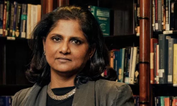 A serious woman in a gray blazer stands in front of a bookshelf filled with books, conveying a professional and academic atmosphere.
