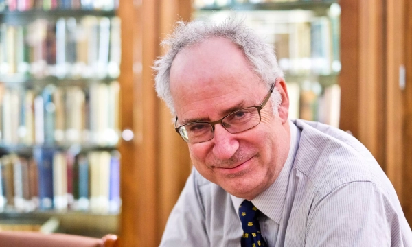 A smiling man with glasses and gray hair sits at a table in a library, surrounded by shelves of books.