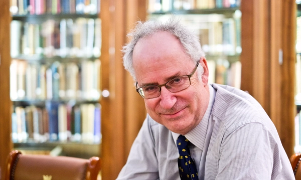 A man with glasses and a gray tie seated in a library with bookshelves in the background.