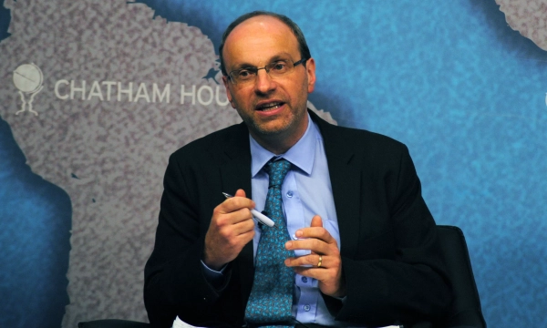 A man in a suit and tie speaks while seated, gesturing with a pen, in front of a map of South America and the Chatham House logo.