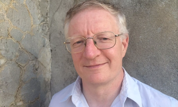 A middle-aged man with glasses stands in front of a textured gray wall, wearing a light-colored shirt and smiling softly.