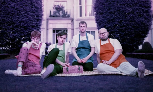 Four individuals sit on a picnic blanket in a garden, wearing colorful aprons. They appear relaxed, surrounded by manicured hedges and a stately building in the background.