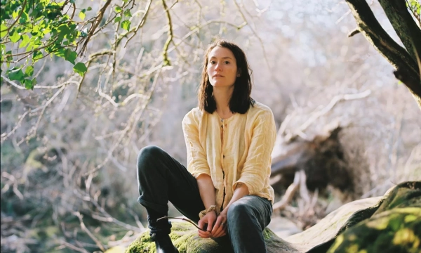A young individual sits on a moss-covered log in a sunlit forest, surrounded by trees and soft foliage, evoking a serene, peaceful atmosphere.