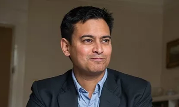 A man in a black blazer and blue shirt smiling slightly while sitting indoors, with a neutral background.