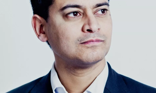A serious-looking man in a dark suit with a light-colored shirt, gazing thoughtfully into the distance against a plain background.