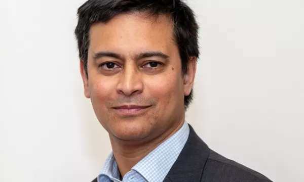 A man in a dark suit and light blue shirt poses against a plain white background, displaying a confident and professional demeanor.
