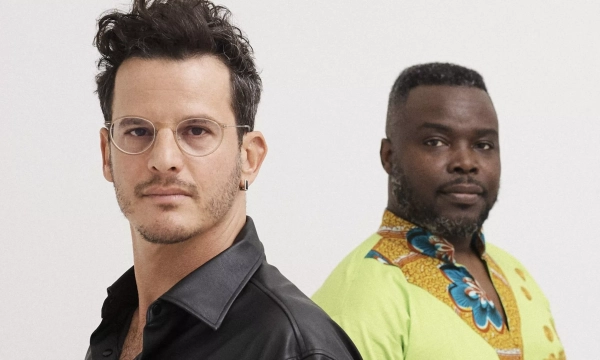 Two men stand back-to-back, one in a black shirt with glasses and the other in a vibrant green shirt with colorful patterns.