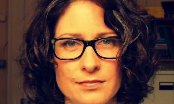 A woman with curly hair and glasses is looking directly at the camera, wearing a striped shirt, with an indoor setting in the background.