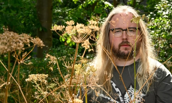 Man with long hair and glasses sits amidst dried plants in a lush green garden.