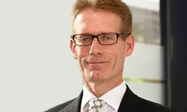A professional man with glasses and a suit, smiling slightly, in a modern office setting with natural light.