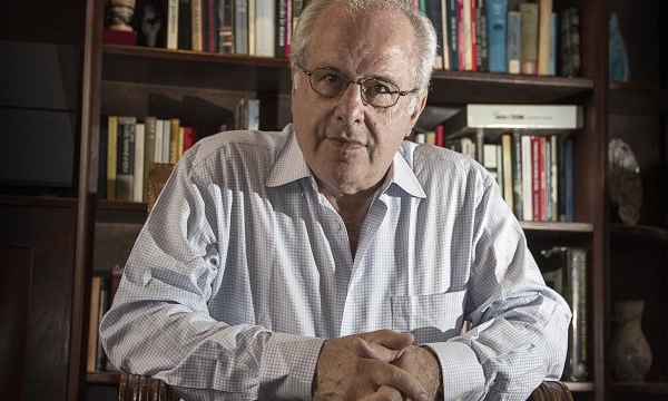 A middle-aged man with glasses sits in front of a bookshelf filled with books, wearing a light checkered shirt and looking directly at the camera.