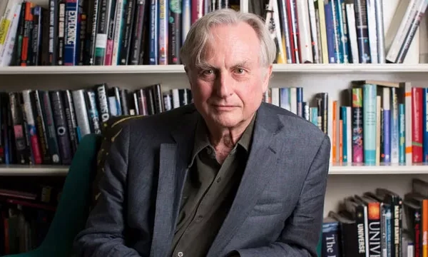 A middle-aged man with silver hair sits in front of a bookshelf filled with various books, wearing a gray blazer and a dark shirt.