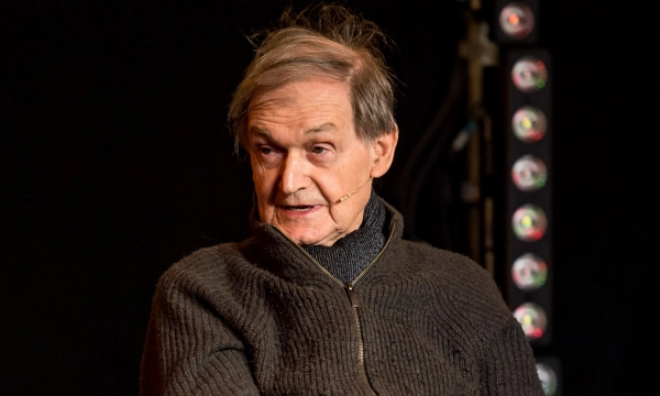 A middle-aged man with grey hair and a microphone headset speaks during an event, wearing a dark sweater in front of a dimly lit background.