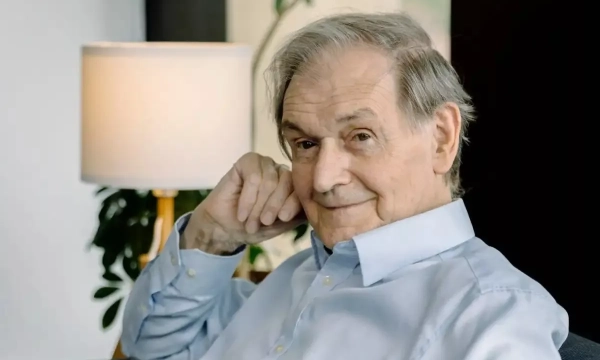 An older man with gray hair and a gentle smile sits thoughtfully in a modern living space, with a lamp and plants in the background.