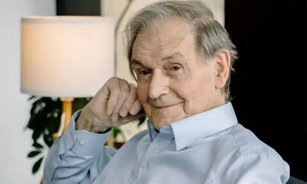 An elderly man with gray hair and a gentle smile, wearing a light blue shirt, seated in a cozy environment with a lamp and plants in the background.