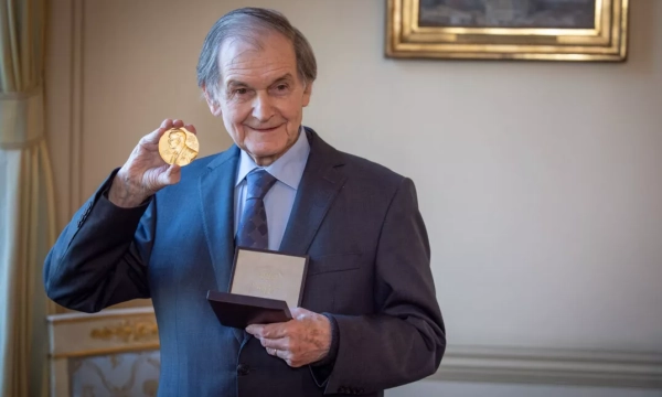 A distinguished elderly man in a suit holds up a Nobel Prize medal, smiling proudly in a formal indoor setting.