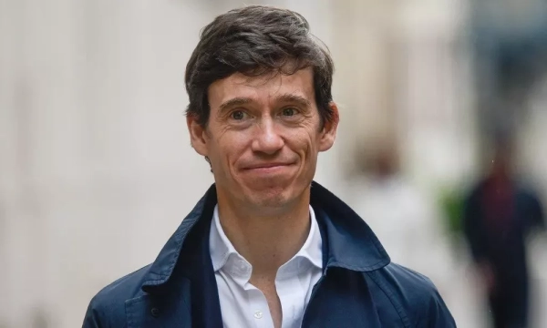 A man in a dark jacket and white shirt smiles while walking outdoors, with a blurred background of people and cityscape.