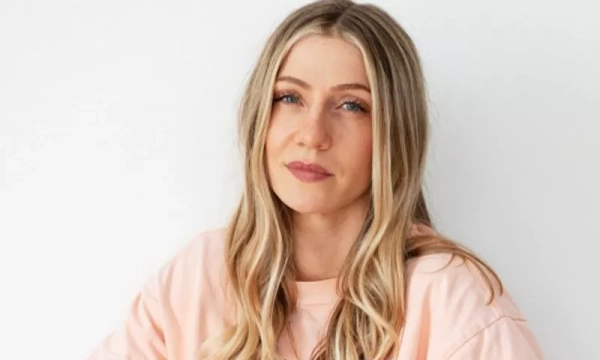 A woman with long, wavy blonde hair wearing a light pink sweatshirt, standing against a plain white background with a serious expression.