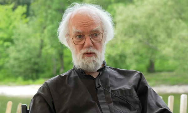 An older man with white hair and a beard, wearing glasses and a black shirt, appears thoughtful against a natural green backdrop.
