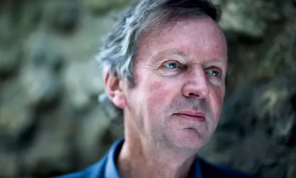 A close-up portrait of a man with gray hair and blue eyes, set against a textured, natural background. He has a thoughtful expression.