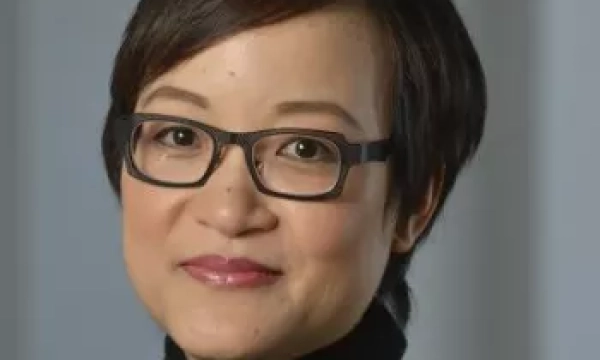 A smiling woman with short dark hair and glasses, wearing a black turtleneck, stands against a blurred background.