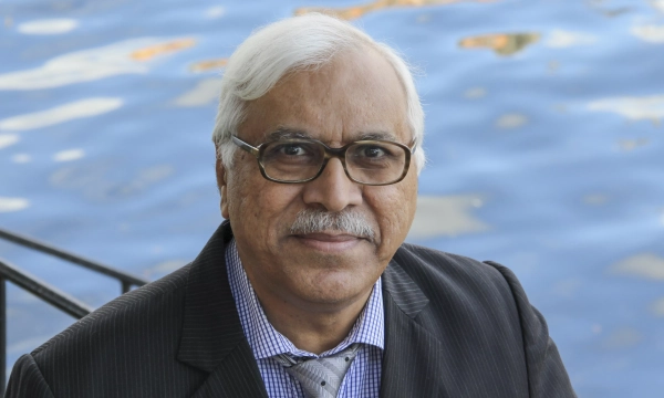 A man with gray hair and glasses, wearing a black suit and tie, smiles while standing by a body of water.
