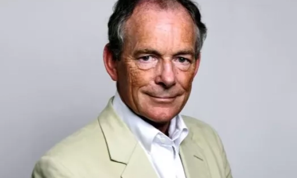 A man in a light-colored blazer and white shirt smiles confidently against a plain background, showcasing a mature appearance.