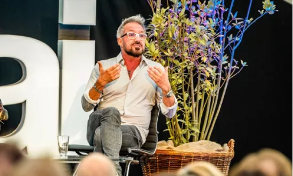 A speaker gestures while seated on a wicker chair, surrounded by plants, during a panel discussion or event.