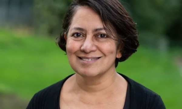 A middle-aged woman with short, dark hair and a warm smile stands outdoors against a green background, wearing a black cardigan.