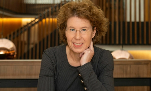 Portrait of a woman with curly hair and glasses, smiling while resting her chin on her hand, set against a modern interior background with wooden elements.
