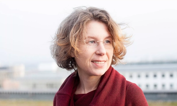 A woman with curly hair and glasses smiles softly while standing outdoors, wearing a maroon sweater against a blurred background.
