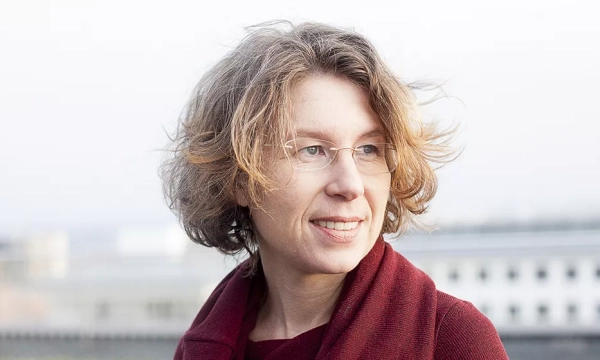 A woman with curly brown hair and glasses smiles softly outdoors, wearing a red sweater and appears to be in a thoughtful pose against a blurred urban backdrop.