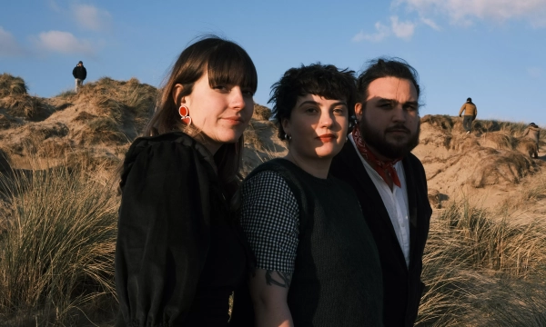 Three individuals stand together on sand dunes, with grass and blue sky in the background. They exhibit a mix of expressions and styles.
